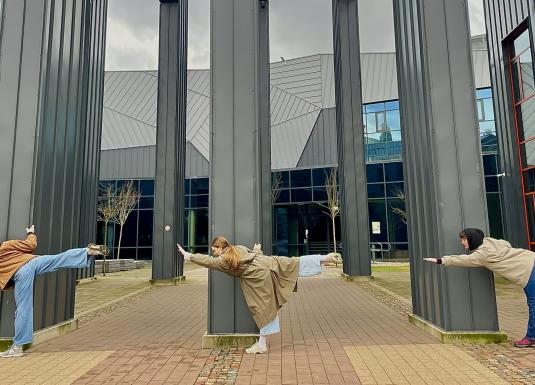 Trīs jaunieši pie muzeja ēkas kolonnām izpilda līdzsvara pozu ar izstieptām kājām un rokām.