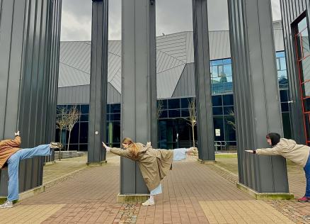 Trīs jaunieši pie muzeja ēkas kolonnām izpilda līdzsvara pozu ar izstieptām kājām un rokām.