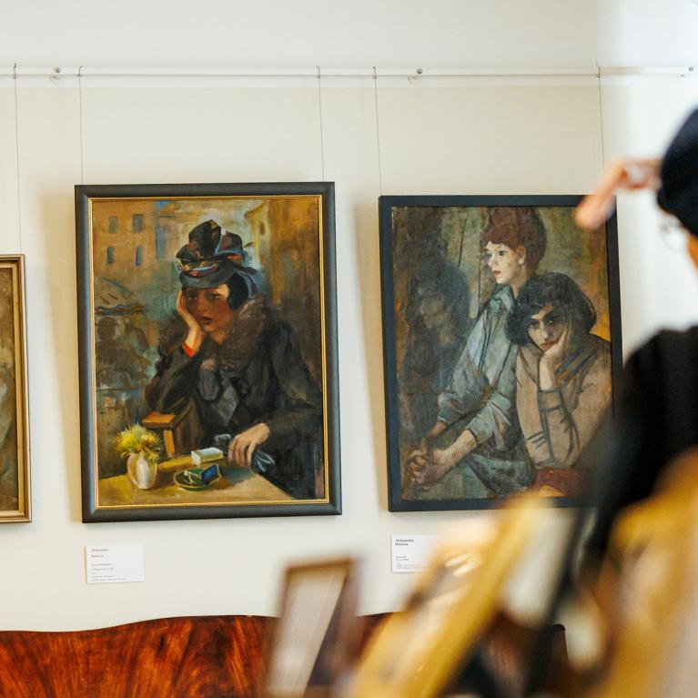 Muzeja sienā izkārtotas trīs gleznas ar cilvēku portretiem, kamēr priekšplānā apmeklētāja žestikulē, skatoties uz darbiem.