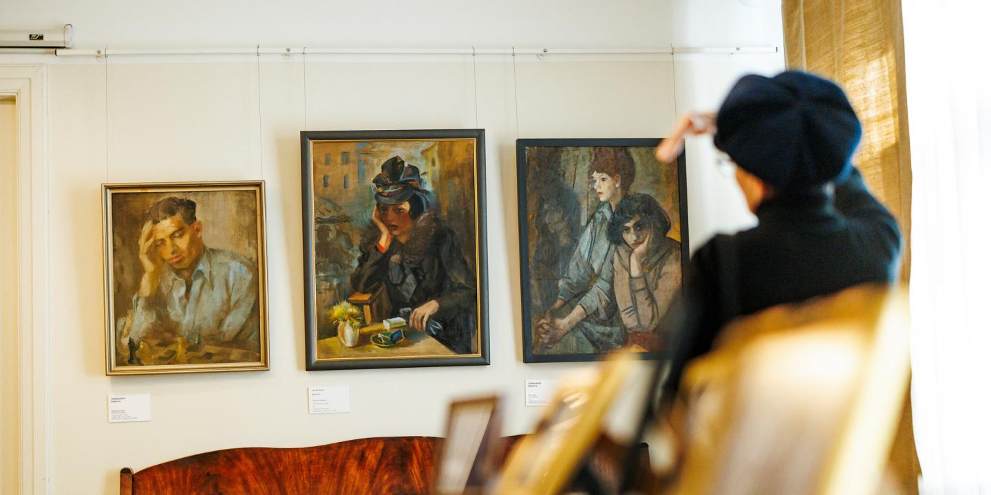 Muzeja sienā izkārtotas trīs gleznas ar cilvēku portretiem, kamēr priekšplānā apmeklētāja žestikulē, skatoties uz darbiem.