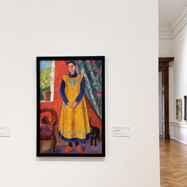 Muzeja interjerā redzama gleznu ekspozīcija uz baltas sienas ar dažādiem darbiem rāmjos.