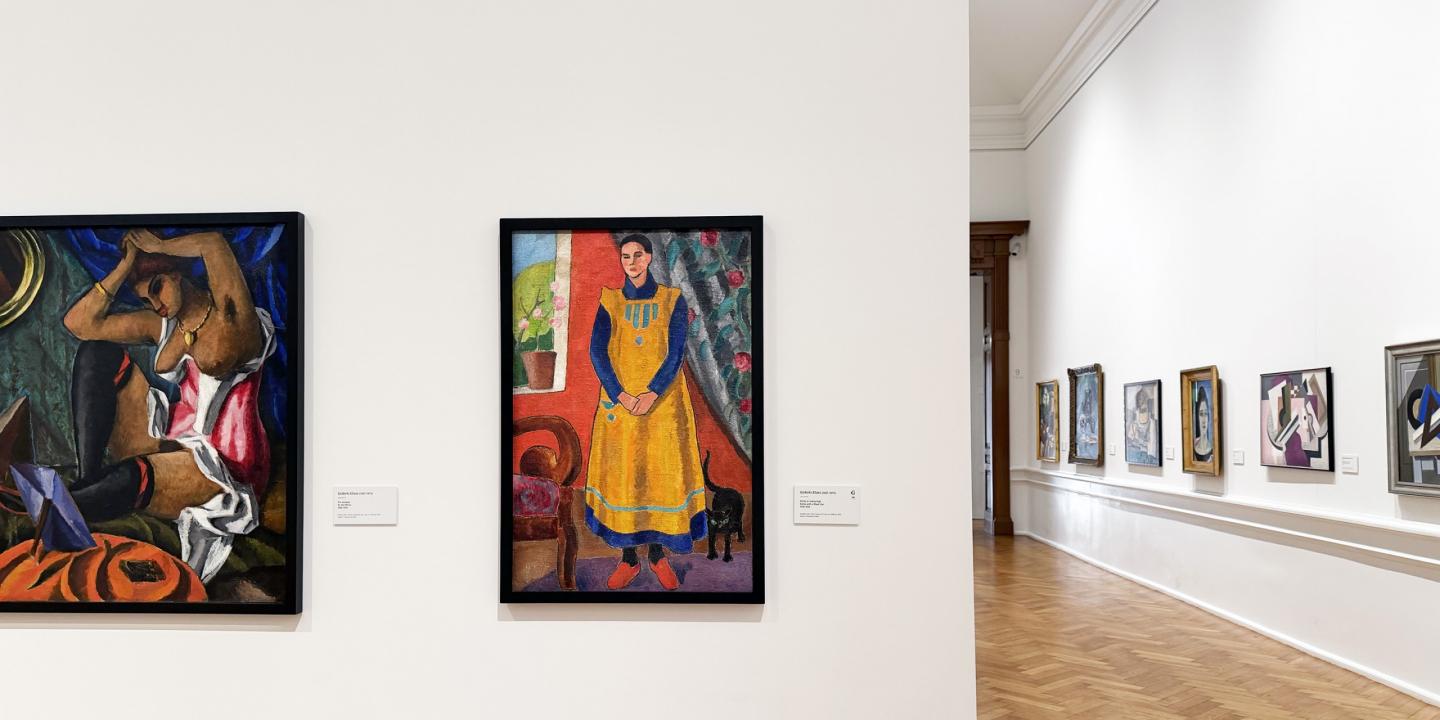 Muzeja interjerā redzama gleznu ekspozīcija uz baltas sienas ar dažādiem darbiem rāmjos.