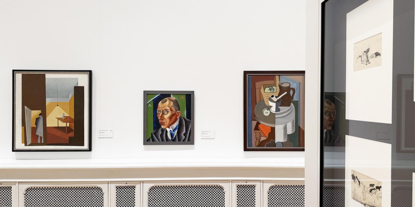 Muzeja zāles sienā izvietotas vairākas gleznas, tostarp portrets un klusā daba, baltā interjerā.