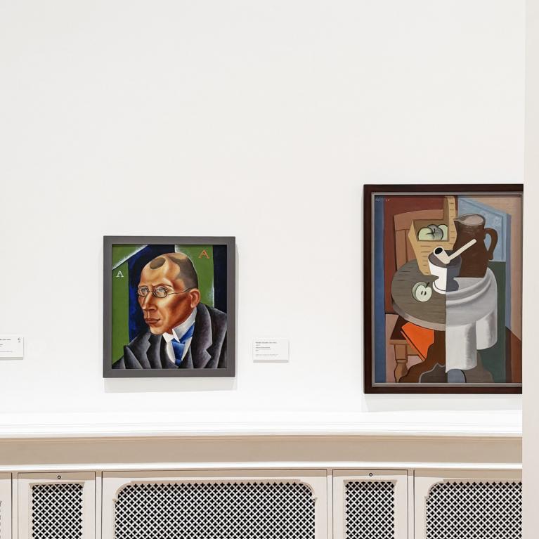 Muzeja zāles sienā izvietotas vairākas gleznas, tostarp portrets un klusā daba, baltā interjerā.