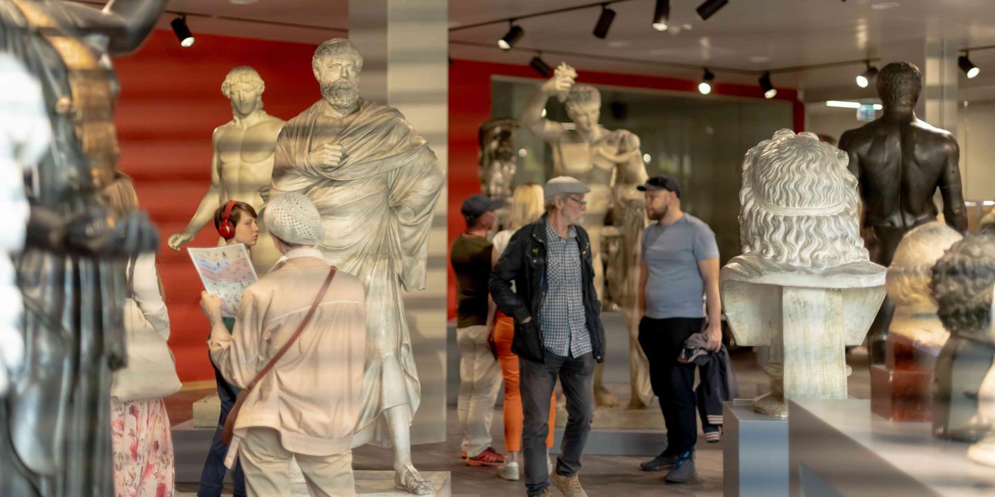 Muzeja zālē ar klasiskām skulptūrām apmeklētāji aplūko eksponātus un lasa informācijas materiālus.