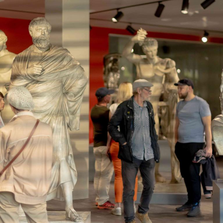 Muzeja zālē ar klasiskām skulptūrām apmeklētāji aplūko eksponātus un lasa informācijas materiālus.