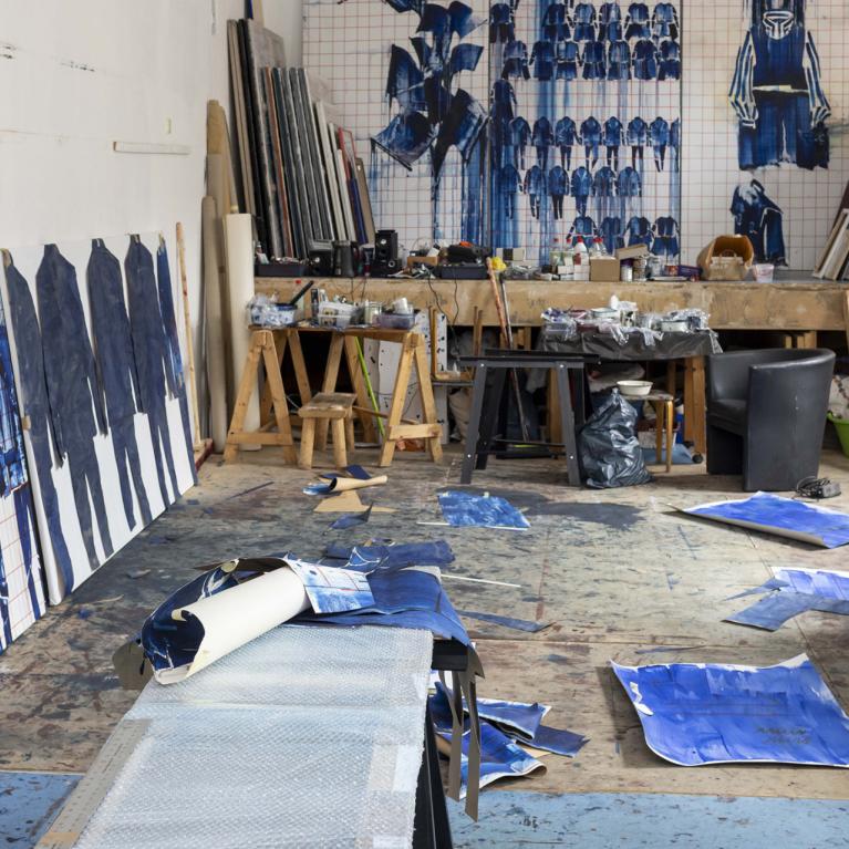 An artist’s studio with large blue paintings and sketches of human figures leaning against the walls and floor, among worktables, tools and canvases.