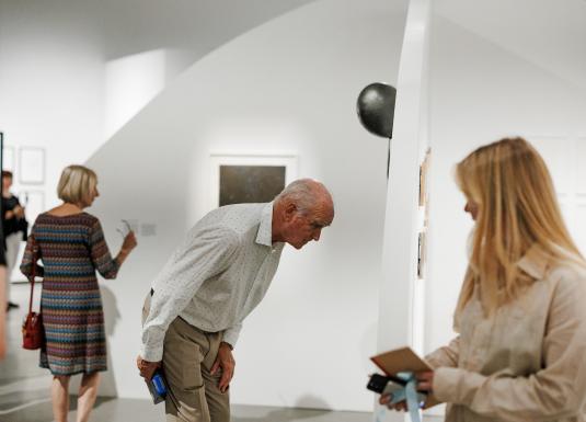 Mākslas galerijas interjerā vecāks vīrietis un vairākas sievietes aplūko pie sienām izvietotus darbus.
