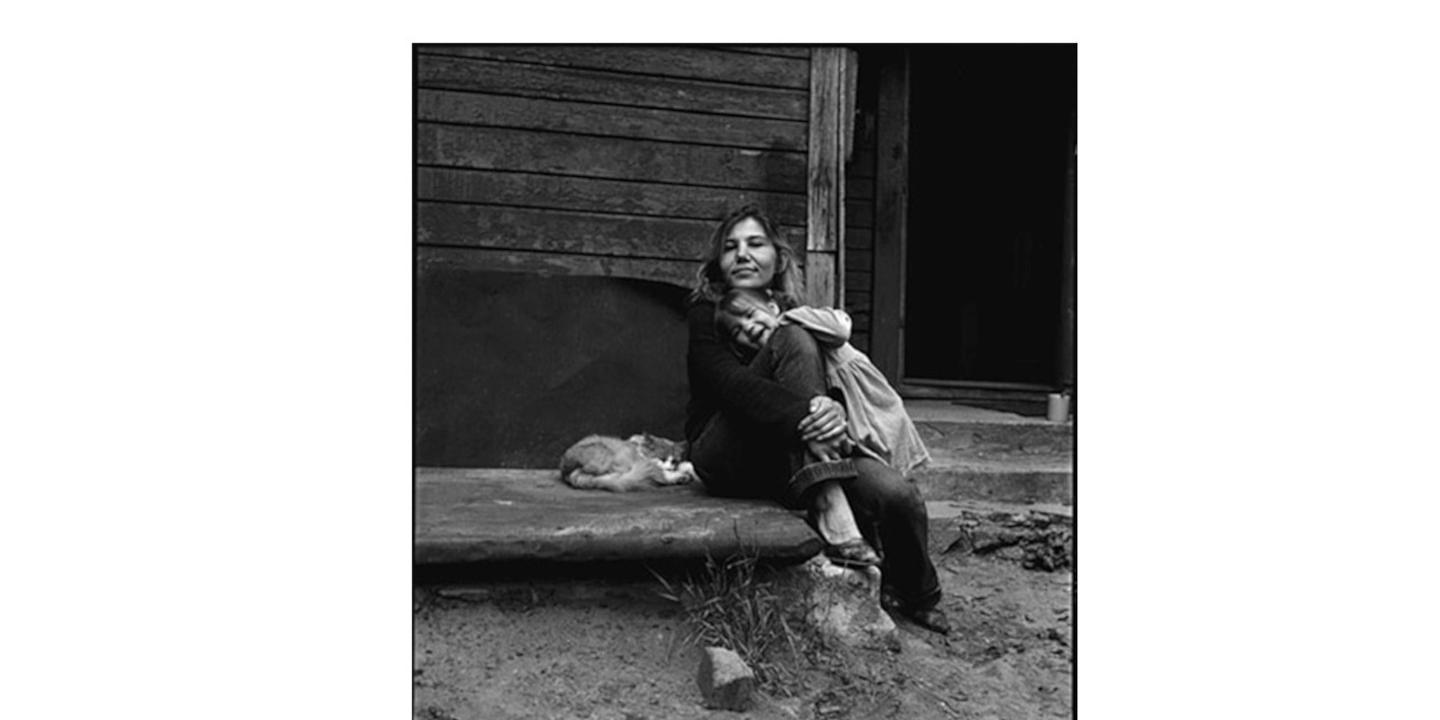 Melnbalta fotogrāfija, kur sieviete sēž uz koka ēkas lieveņa, apskāvusi bērnu, bet blakus uz pakāpiena guļ kaķis.