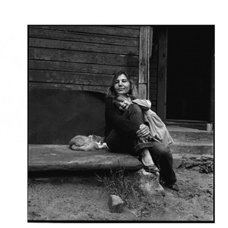 Melnbalta fotogrāfija, kur sieviete sēž uz koka ēkas lieveņa, apskāvusi bērnu, bet blakus uz pakāpiena guļ kaķis.