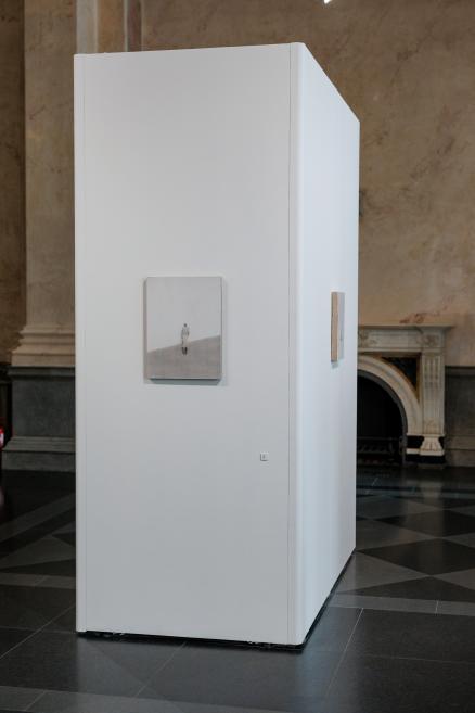 Small-format artworks are displayed on a white exhibition wall in a museum gallery, with historic interior architecture and a fireplace visible in the background.