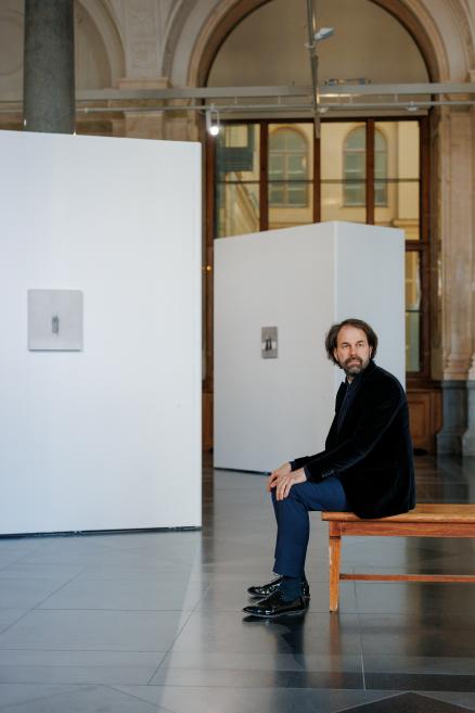 A man sits on a bench in a museum exhibition hall with high arches and white display walls, with exhibited artworks visible in the background.