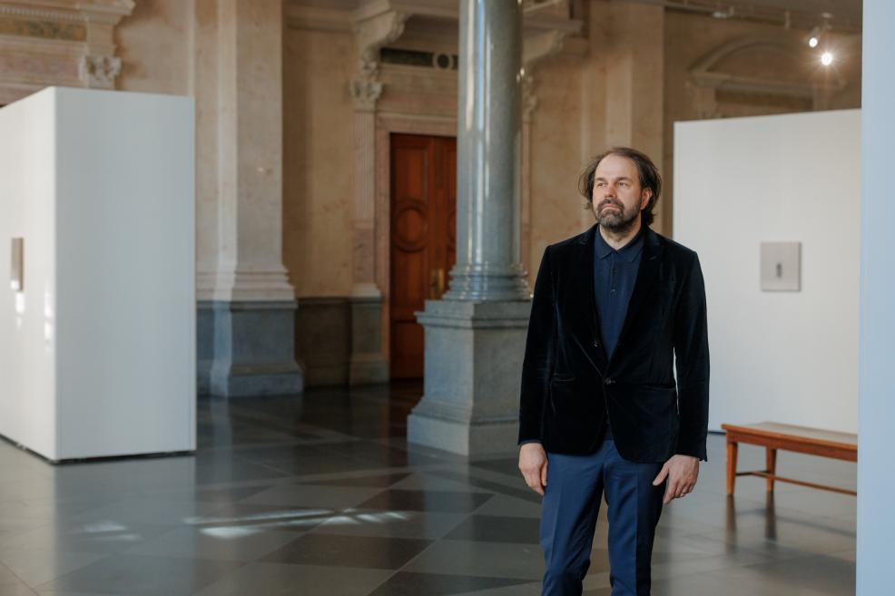 A man stands in a museum exhibition hall among white display walls, historic columns, and a bench, looking toward the artworks installed in the space.