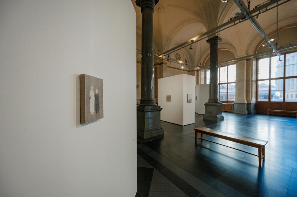 Small-format artworks are displayed on white exhibition walls in a historic museum gallery with tall windows, granite columns, and a bench for visitors.