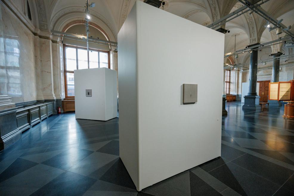 Small-format artworks are displayed on white exhibition walls in a historic museum gallery surrounded by tall windows, decorative vaults, and granite columns.