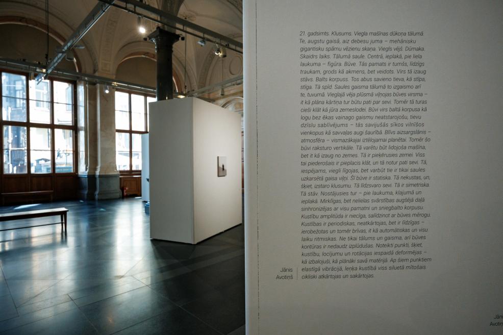 In a historic museum gallery, an artist’s text is displayed on a white exhibition wall, with exhibition structures and small-format artworks visible in the background.