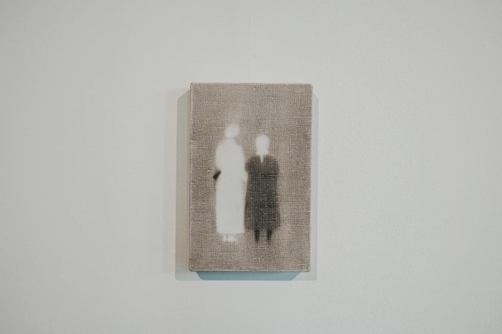 A small-scale artwork depicts two blurred human figures—one light and one dark—against a grey-toned background, creating a subdued and symbolic atmosphere.