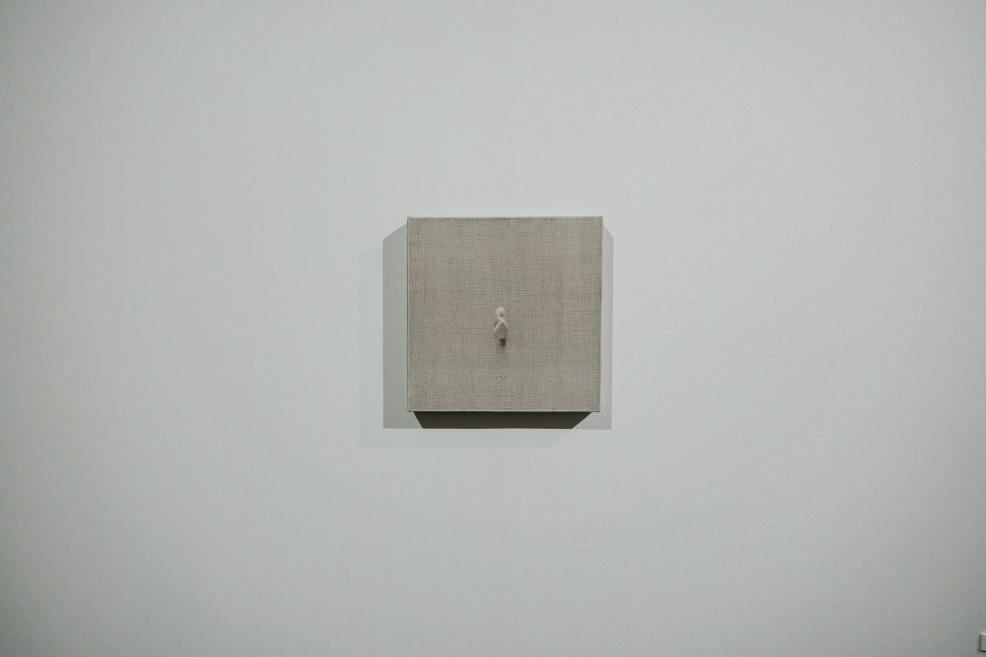 At the center of a small square artwork is a tiny stylized human figure set against a light monochromatic background, creating a minimalist and contemplative atmosphere.