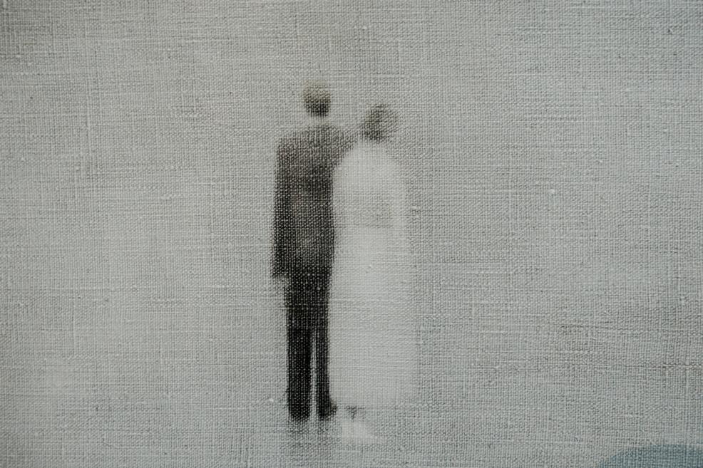 Two blurred human figures, one dark and one light, stand closely together on a grey-toned canvas, creating an impression of fragile intimacy and memory.