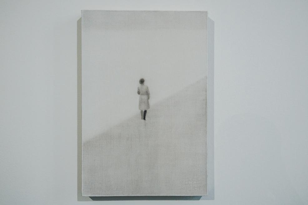 A small, blurred human figure is depicted alone on a gently sloping, barely discernible surface, evoking a sense of silence, solitude, and an uncertain journey.