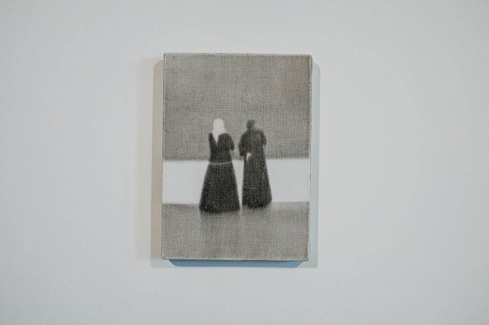 A small rectangular canvas depicts two blurred human figures in dark clothing standing side by side against a grey background with a light horizontal band, evoking a sense of quiet presence, connection, and memory.