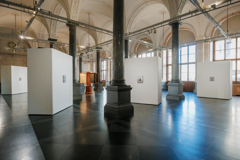 White exhibition walls displaying small-format artworks are arranged among monumental columns in a historic museum gallery, creating a spacious and harmonious exhibition setting.