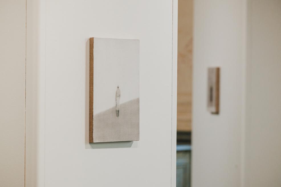 A close-up view of a small-format artwork on a white exhibition wall depicts a solitary human figure in a minimalist, light-toned landscape.