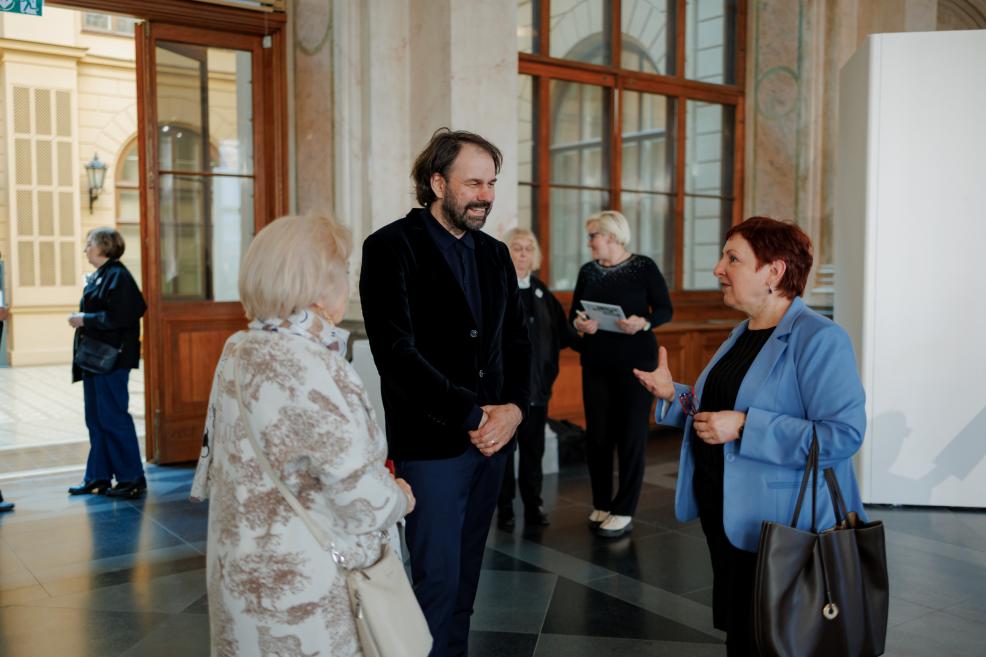 Several visitors are conversing in a historic museum exhibition hall, while in the foreground a man in a dark jacket is engaged in conversation with a woman wearing a blue blazer.