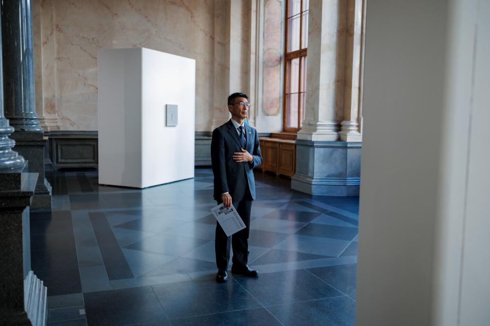 In a historic museum exhibition hall, a man in a suit stands near display walls featuring small-format artworks while holding exhibition materials in his hand.