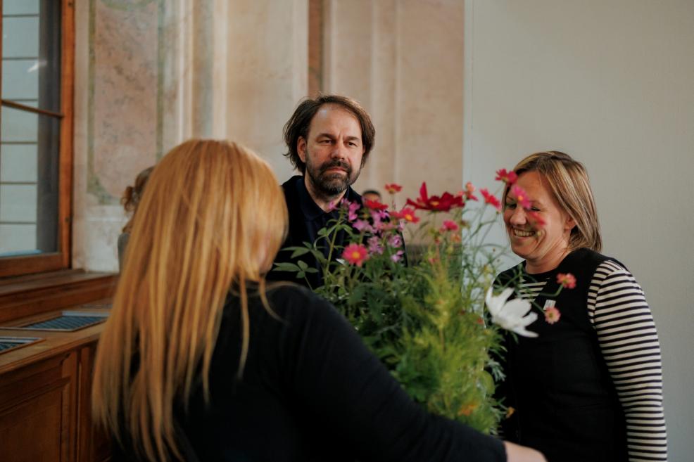 People converse beside a bouquet of colourful flowers during a museum exhibition opening in a historic gallery interior.