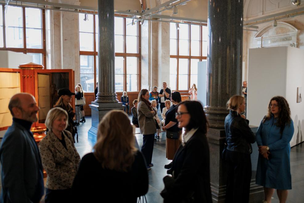 Visitors gather and converse at a museum exhibition opening in a spacious historic hall with tall windows, columns, and exhibition displays.