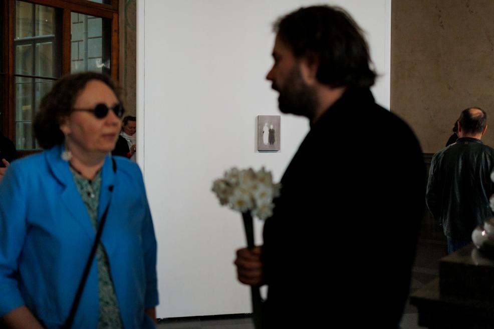 Two exhibition visitors converse in a museum gallery beside a white display wall featuring a small artwork, while one of them holds a bouquet of white flowers.