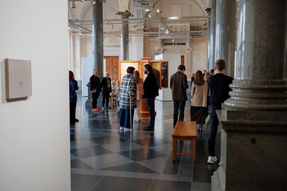 Visitors explore the exhibition in a spacious museum gallery with tall columns, white display walls, and small-scale artworks, gathering to converse and observe the works.