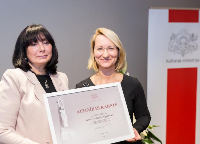 Divas sievietes pasākumā tur Kultūras ministrijas atzinības rakstu, fonā redzams Kultūras ministrijas baneris.