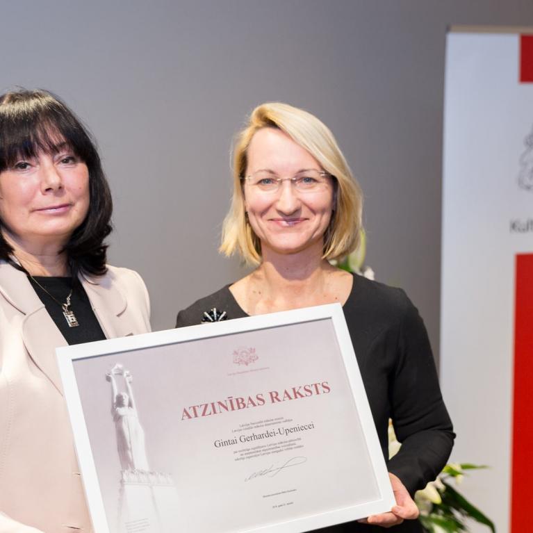 Divas sievietes pasākumā tur Kultūras ministrijas atzinības rakstu, fonā redzams Kultūras ministrijas baneris.