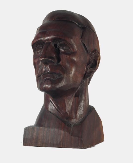 A dark wooden sculpture depicts a male bust with a stylized face against a white background.