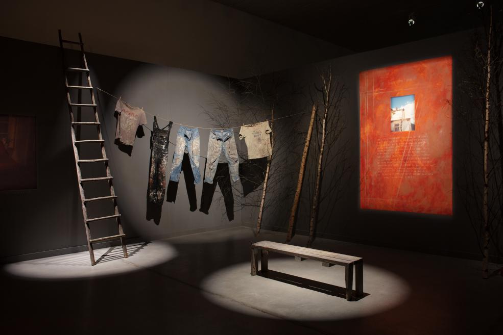 Museum installation with a wooden ladder, worn clothes hanging on a line, a wooden bench, and a red wall work with a photographic inset.