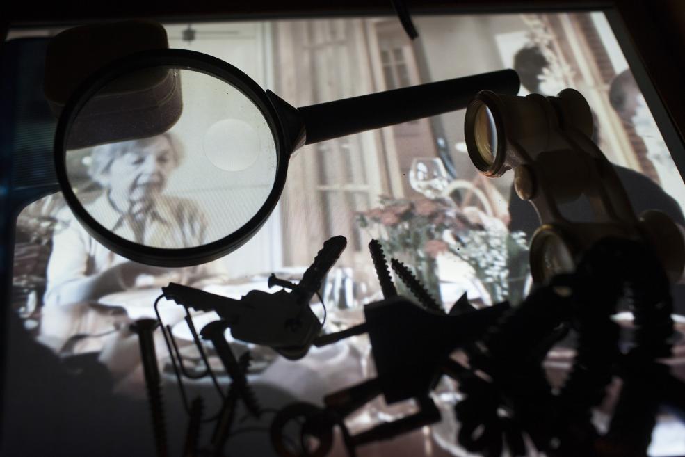 Dark installation view with a magnifying glass and binoculars in the foreground framing a blurred video projection of a seated woman at a table.