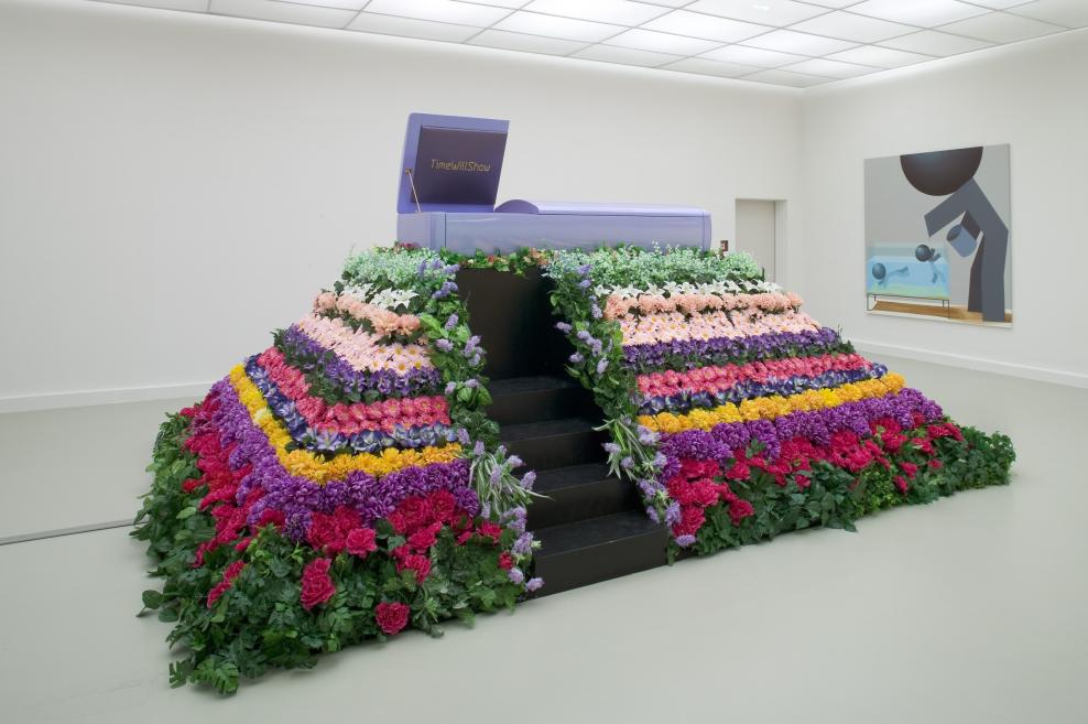 Balta galerijas telpa ar violetu instalāciju kā sarkofāgu vai kapsulu, ko ieskauj krāsainu ziedu slāņi un melnas kāpnes priekšpusē, pie sienas redzama abstrakta glezna.