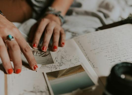 Tuvplānā redzamas personas rokas ar sarkani lakotiem nagiem, kas pārlūko pierakstus, skices un fotogrāfijas uz gultas, radot radoša darba un pārdomu noskaņu.