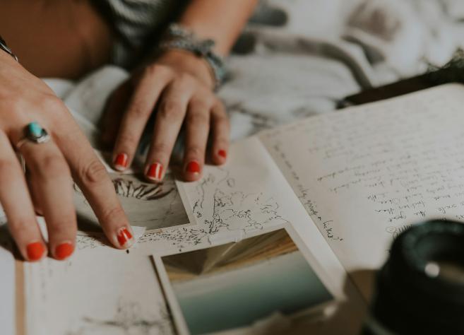 Tuvplānā redzamas personas rokas ar sarkani lakotiem nagiem, kas pārlūko pierakstus, skices un fotogrāfijas uz gultas, radot radoša darba un pārdomu noskaņu.
