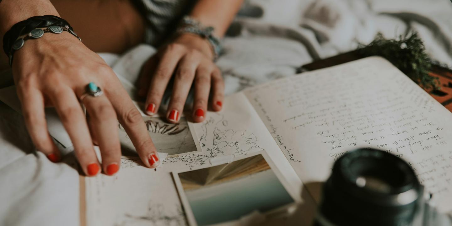 Tuvplānā redzamas personas rokas ar sarkani lakotiem nagiem, kas pārlūko pierakstus, skices un fotogrāfijas uz gultas, radot radoša darba un pārdomu noskaņu.