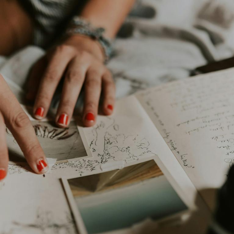 Tuvplānā redzamas personas rokas ar sarkani lakotiem nagiem, kas pārlūko pierakstus, skices un fotogrāfijas uz gultas, radot radoša darba un pārdomu noskaņu.