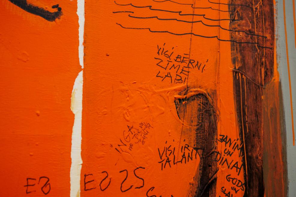 Oranžas gleznas fragmentā redzami melnā krāsā uzrakstīti teksti un zīmējumi.