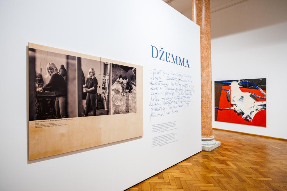 Izstādes skatā redzamas vēsturiskas fotogrāfijas ar mākslinieci un uzraksts "Džemma", kā arī liela abstrakta glezna uz sarkanā fona.