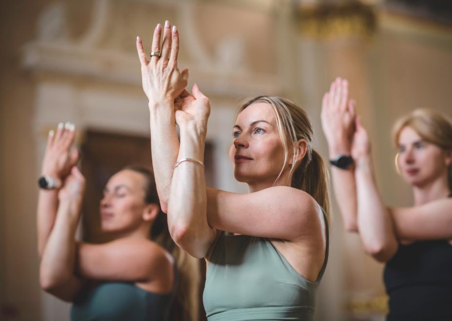 Sievietes grupā izpilda jogas elpošanas un roku pacelšanas vingrinājumu klasiskā interjerā.