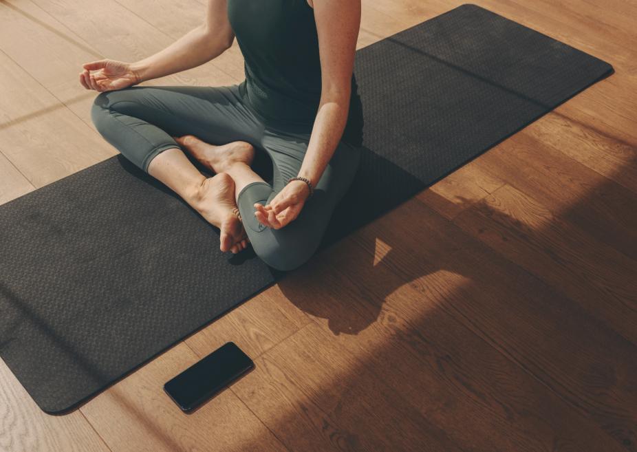 Persona sēž meditācijas pozā uz jogas paklāja koka grīdas telpā.