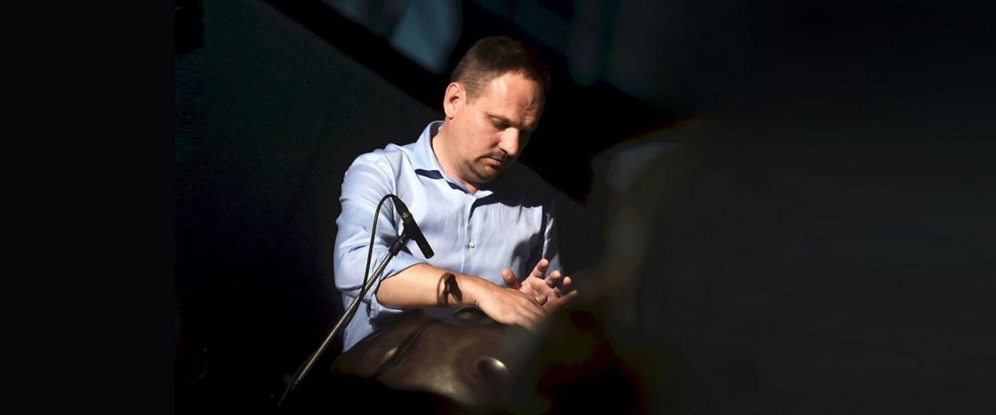 Vīrietis elegantā tumšā uzvalkā stāv pie tumšas sienas ar rokām sakrustotām un skatās sāņus, gaismā veidojot asu ēnu.