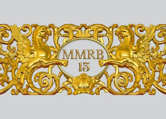Zeltīts dekoratīvs ornaments ar diviem spārnotiem grifiem, vīnogu ķekariem un centrālu medaljonu ar uzrakstu “MMRB 15”.