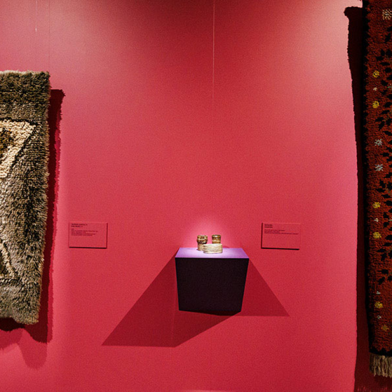 Exhibition view with textile artworks displayed on a dark pink wall: two narrow greyish rugs with geometric patterns and a large red-orange rug with an ornamental border.